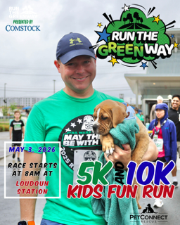 Man in a green shirt holding a brown dog in front of a Run the Green Way promo banner for a 5K/10K Kids Fun Run, May 3, 2026 at Loudoun Station.
